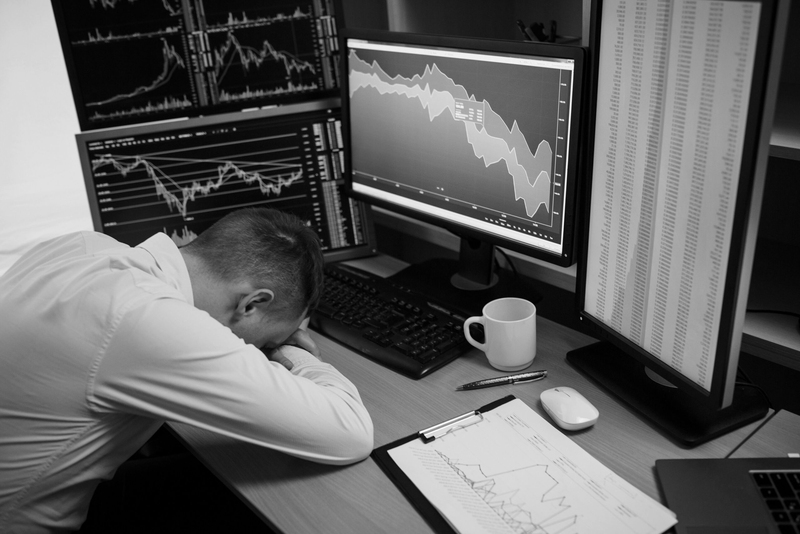 A man sleeps at his desk, resting his head on his arms, with two computer monitors glowing in the background