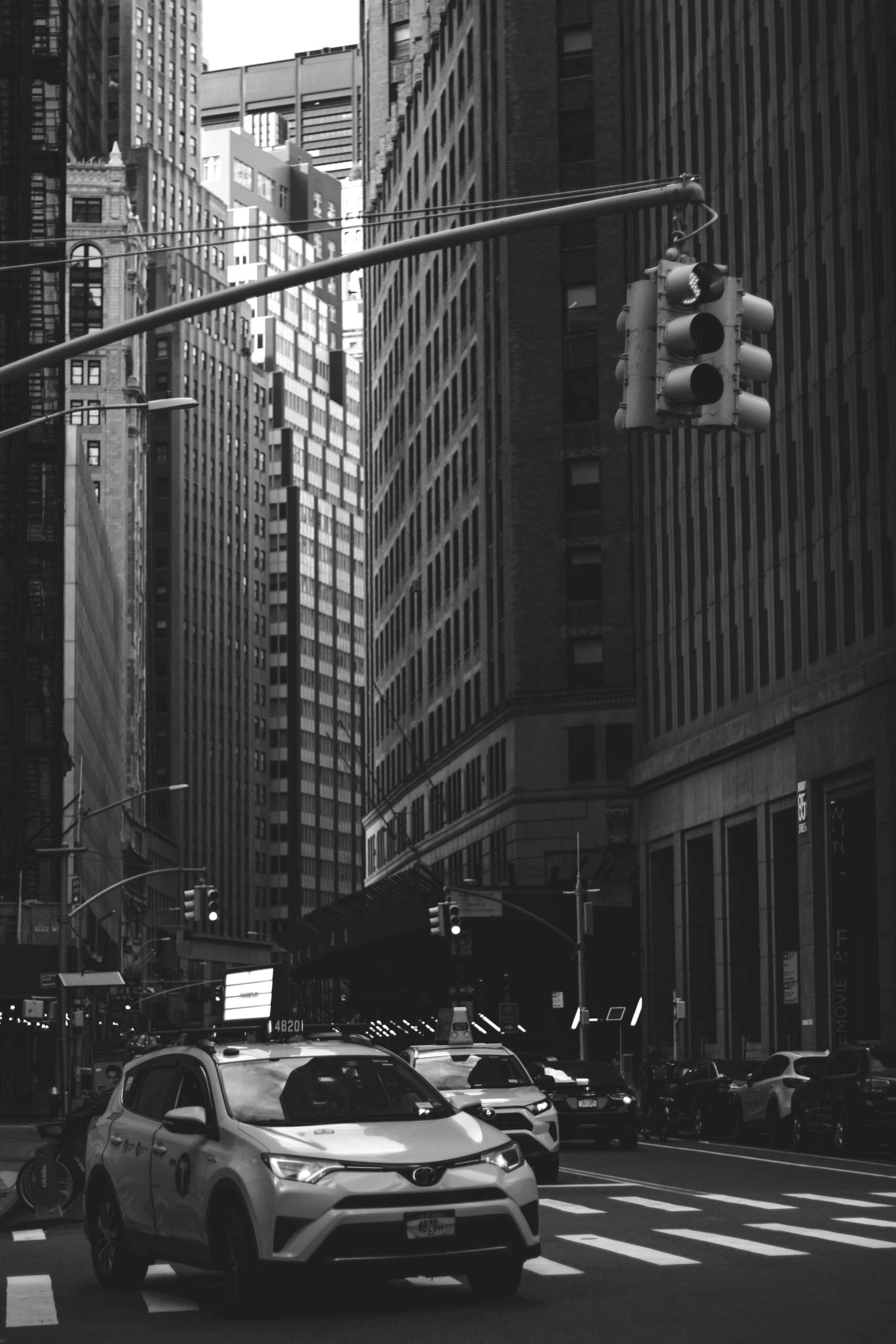 A black and white photo of a car driving along a city street, surrounded by buildings